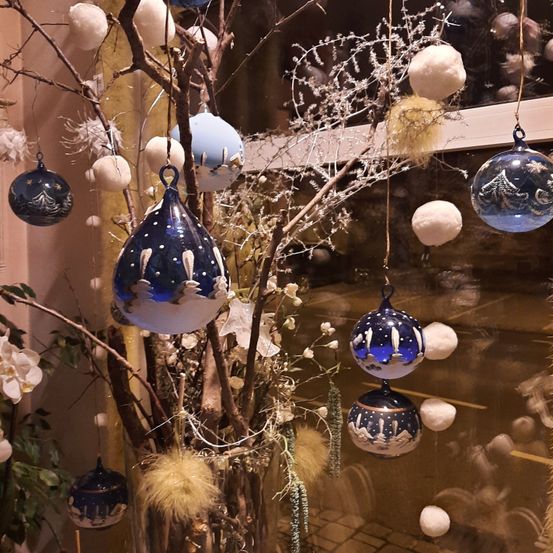 Eine Ausstellung von blauen und weißen Weihnachtskugeln, die von Zweigen mit weißen Blumen und Eiszapfen hängen. Die Kugeln sind aus Glas und mit Schneeszenen verziert.