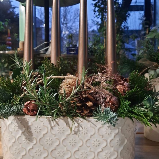 Ein weißer Keramiktopf mit einem dekorativen Muster enthält eine festliche Anordnung aus Grünpflanzen, Tannenzapfen und Rosmarin, akzentuiert durch hohe bronzefarbene Kerzenhalter. Der Innenraum ist mit einem verschwommenen Blick auf einen glasverkleideten Raum und Bäume draußen zu sehen.