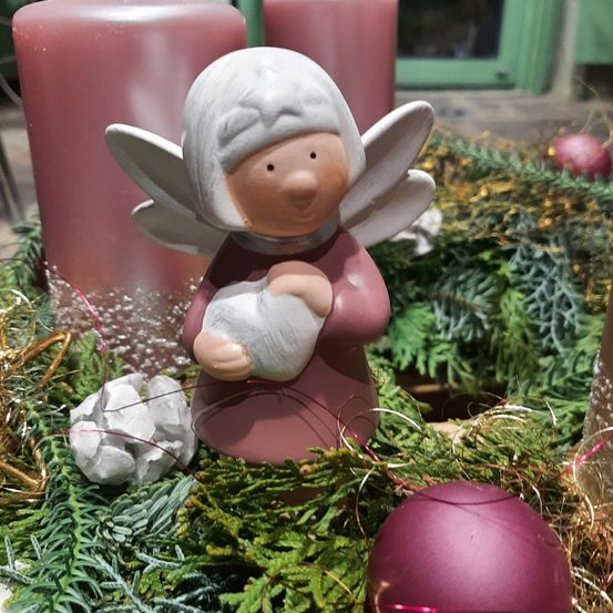 Eine kleine Engelfigur wiegt ein Baby und ist von Weihnachtsdekorationen umgeben, einschließlich einer Girlande, Kerzen und Ornamenten, auf einer Holzoberfläche.