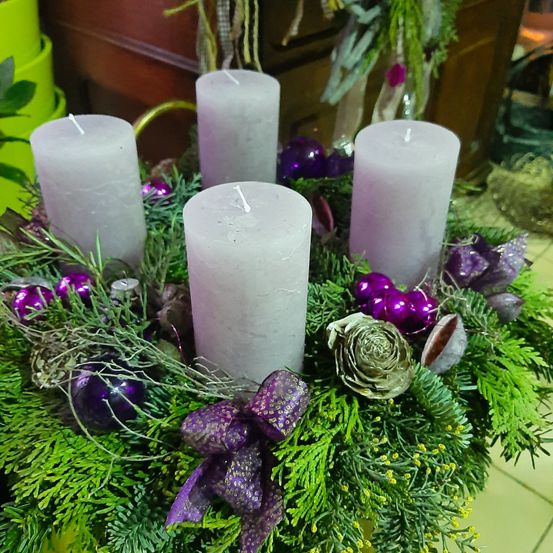 Ein Kranz mit vier weißen Kerzen, lila Ornamenten und grünen Blättern. Die Kerzen sind nicht angezündet, und der Kranz ist mit einem lila Band dekoriert.
