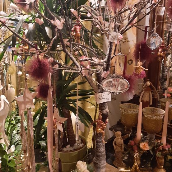 Ein dekorativer Baum, geschmückt mit rosa Ornamenten und kleinen Figuren, umgeben von verschiedenen Pflanzen und Objekten. Der Baum hat Äste mit Ornamenten und steht neben einer Kerze und einer Figur auf einem Tisch.