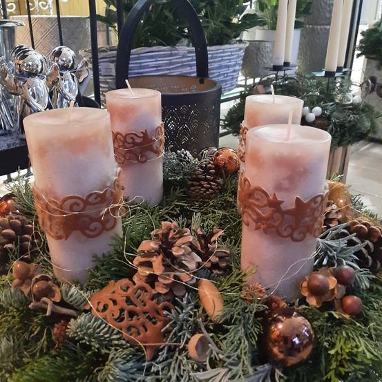 Vier rosa Kerzen mit dekorativen Mustern sind in einem Kranz mit Tannenzapfen und Weihnachtsdekorationen angeordnet. Hinter dem Kranz befinden sich mehrere Kerzen und ein Korb mit einer Pflanze.