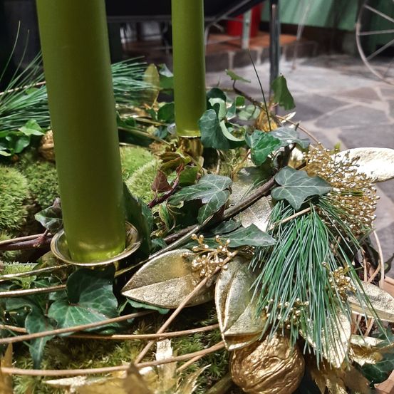 Zwei hohe grüne Kerzen stehen inmitten einer Sammlung von grünen Blättern, goldenen Blättern und Moos. Die Kerzen werden von goldenen Kerzenständern gehalten. Kleine goldene Blätter sind über die Szene verstreut.