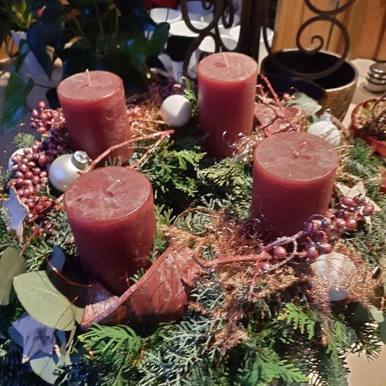 Ein Kranz mit vier roten Kerzen, umgeben von immergrünen Blättern, Beeren und Ornamenten. Die Kerzen sind angezündet, und der Kranz steht auf einem Tisch.