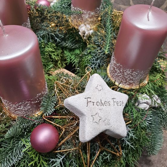 Ein Kranz mit vier rosa Kerzen, einem dekorativen Stern mit dem Text 'Frohes Fest' und einer lila Kugel auf einer Holzoberfläche.