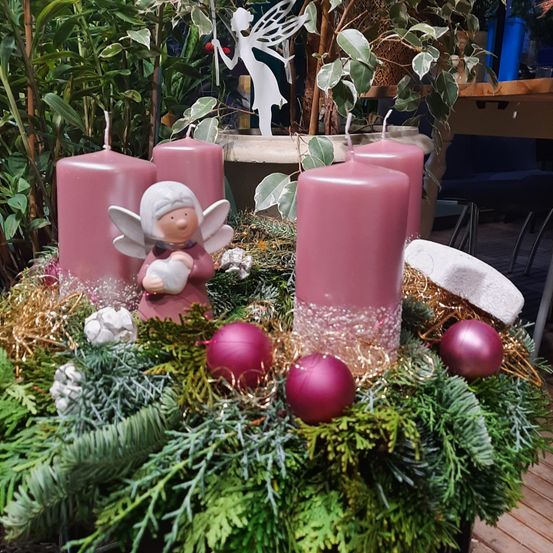 Ein Weihnachtskranz mit rosa Kerzen und Ornamenten, einschließlich einer rosa Engelfigur, steht auf einer Holzoberfläche mit Pflanzen im Hintergrund.