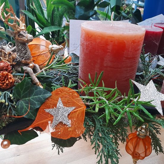 Auf einem Tisch steht eine dekorative Girlande mit einer roten Kerze, einem silbernen Stern und Herbstblättern. Daneben befindet sich eine Figur eines Rentiers.