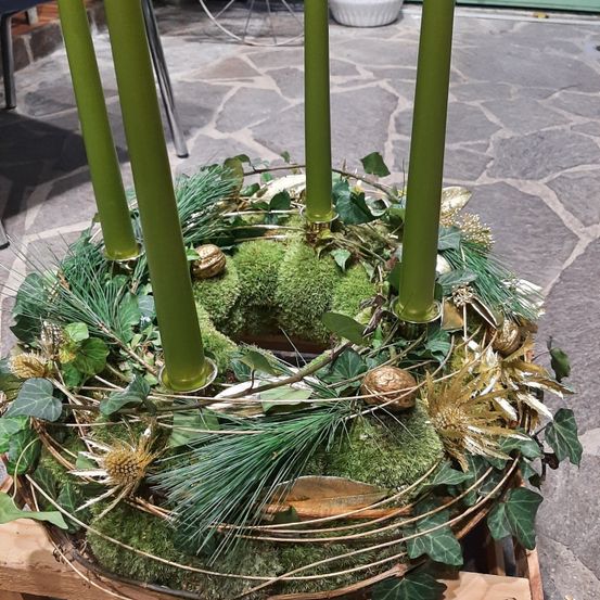 Ein runder Kranz mit grünen Kerzen und goldenen Verzierungen auf einer Holzbasis, umgeben von Moos, Blättern und Zweigen.