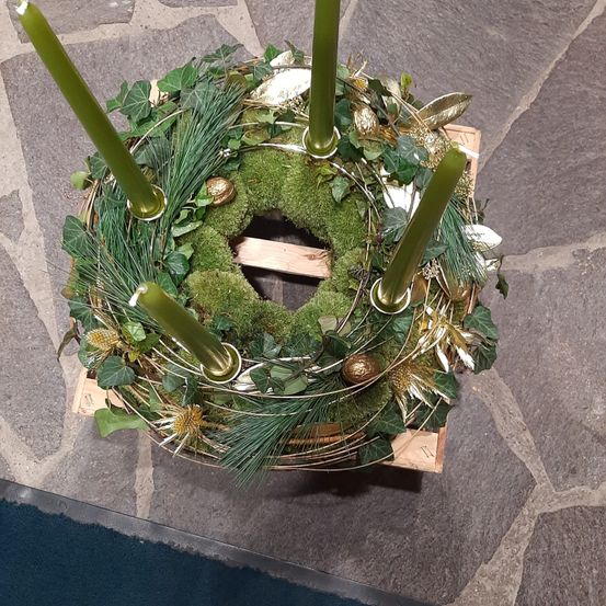 Ein kreisförmiger Kranz mit grünem Moos, Blättern und goldenen Akzenten ruht auf einer Holzbasis mit vier grünen Kerzen.