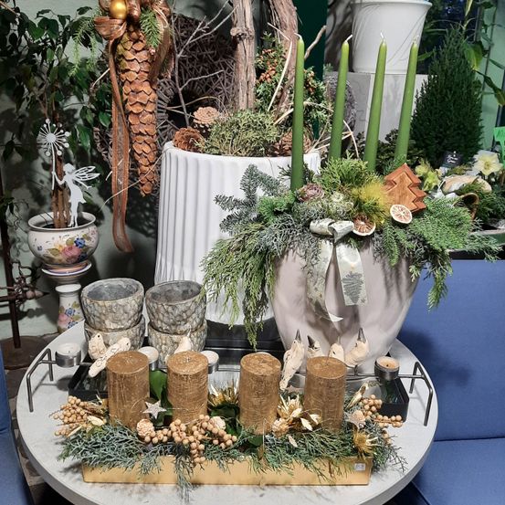 Eine festliche Tischdekoration umfasst eine große Vase mit vier grünen Kerzen, umgeben von Pflanzen, einem dekorativen Vogel und einem Holzbaum. Im Vordergrund stehen fünf goldene Kerzen auf einem Tablett mit verschiedenen Dekorationen.