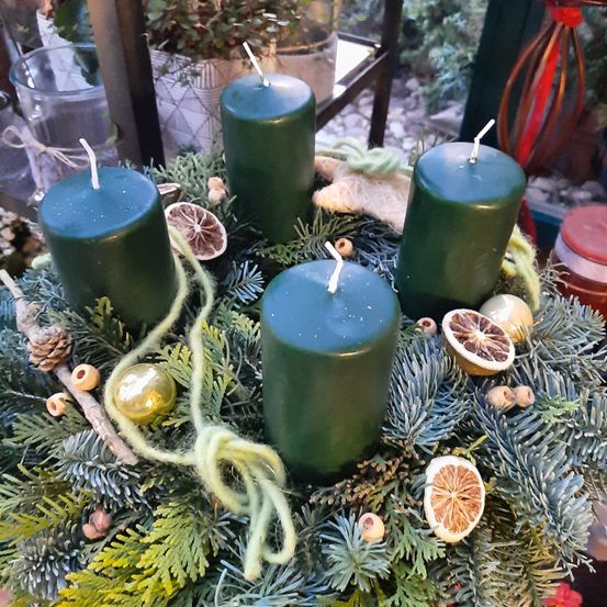 Eine kreisförmige Anordnung mit grünen Kerzen, Tannennadeln und getrockneten Früchten. Es enthält vier grüne Kerzen mit weißen Dochten. Die Kerzen sind von einer Schnur, Tannennadeln und getrockneten Früchten umgeben.