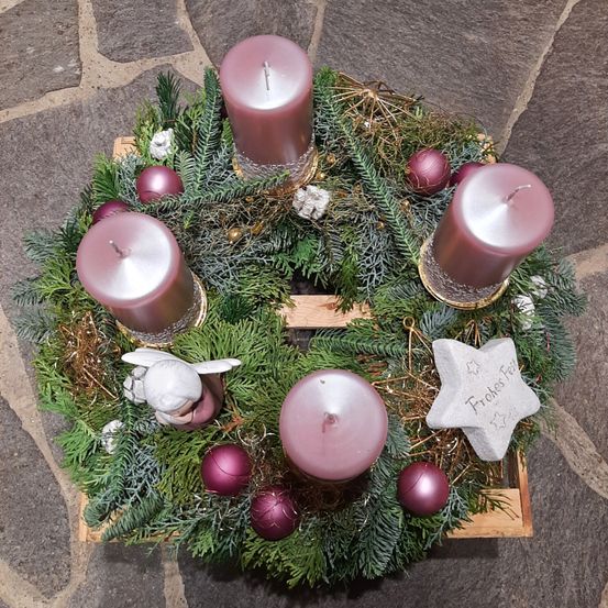Ein Kranz aus Tannennadeln und Ornamenten mit vier rosa Kerzen und einem weißen Engel. Der Kranz liegt auf einer Holzoberfläche, mit einem sternförmigen Ornament mit dem Wort 'Frohe' darauf.