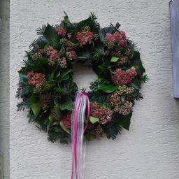 Ein Kranz aus grünen Blättern und rosa Blüten ist an einer weißen Wand befestigt. Es hat ein rosa Band, das herabhängt.