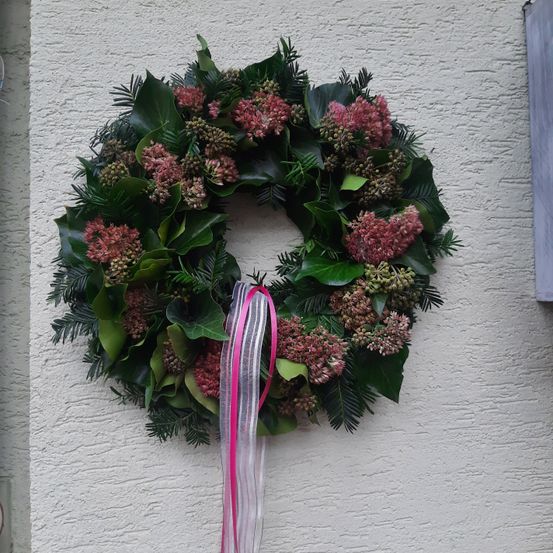 Ein Kranz aus grünen Blättern und rosa Blüten ist an einer weißen Wand befestigt. Es hat ein rosa Band, das herabhängt.