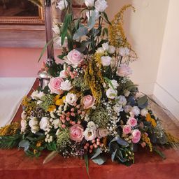 Eine große Blumenarrangement mit weißen, rosa und gelben Blumen, umgeben von grünen Blättern. Es steht auf einem braunen Tisch in einem Raum mit einer rosa Wand und einem gerahmten Bild.