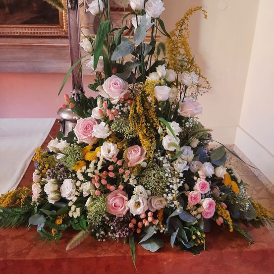 Eine große Blumenarrangement mit weißen, rosa und gelben Blumen, umgeben von grünen Blättern. Es steht auf einem braunen Tisch in einem Raum mit einer rosa Wand und einem gerahmten Bild.