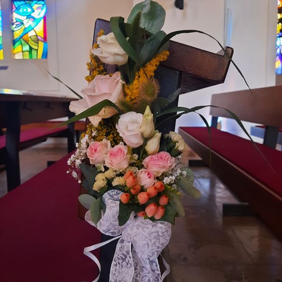 Ein hölzerner Stuhl mit einer Blumenarrangement aus rosa Rosen, gelben Blumen und grünen Blättern steht in einem Kirchengang. Dahinter sind rote Kirchenbänke und Buntglasfenster zu sehen.