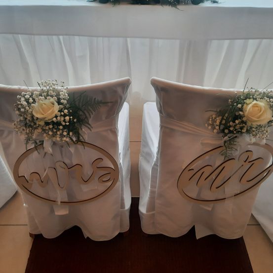 Zwei weiße Stühle sind mit Blumenarrangements geschmückt. Jeder Stuhl hat ein großes Monogramm 'Mr.' und 'Mrs.' auf der Rückseite, mit einem Strauß weißer Blumen und Babys Atmung. Die Stühle sind mit weißem Stoff bezogen, und der Hintergrund ist verschwommen.