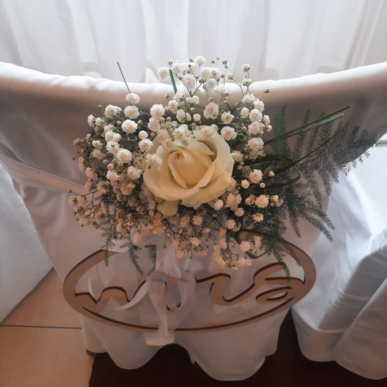 Ein Hochzeitsstuhl geschmückt mit einem Blumenarrangement aus weißen Rosen, Babys Atem und grünen Blättern. Der Stuhl ist mit einem kreisförmigen Schild mit dem Wort 'Viva' versehen.