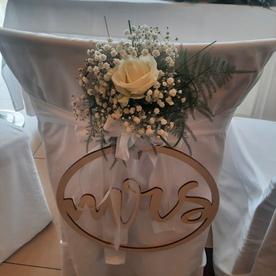 Ein weißer Stuhl, der mit einem weißen Tuch bedeckt ist, geschmückt mit einem Strauß weißer Blumen und einem Schild, das 'Mrs' steht. Der Stuhl ist Teil einer Hochzeitseinrichtung.