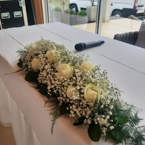 Ein langer weißer Tisch, der mit weißem Stoff bedeckt ist und mit weißen Rosen und Grün bepflanzt ist. Ein schwarzes Mikrofon liegt auf dem Tisch. Im Hintergrund ist ein weißer Van zu sehen.