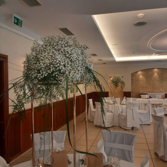 Ein Hochzeitsempfangsbereich ist mit weißen Blumen, weißen Stühlen und weißen Tischdecken dekoriert. Eine große Blumenarrangement ist in der Mitte positioniert, mit zusätzlichen kleineren im ganzen Raum.