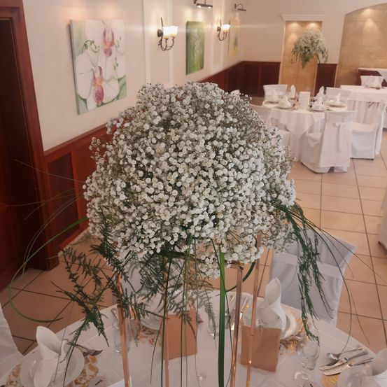 Ein Hochzeitsempfangsort mit weißen Tischdecken und Stühlen, mit einem großen Mittelpunkt aus weißen Babys-Atem-Blumen. Die Wände haben Kunstgemälde, und der Boden ist gefliest.