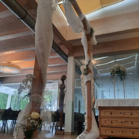 Ein Hochzeitszeremonie-Setup mit einem hölzernen Bogen, geschmückt mit weißem Stoff und Blumen. Der Bereich ist elegant mit weißem Stoff dekoriert, der von der Decke herabhängt. Es gibt zwei Blumenarrangements, eines auf einem Tisch, der mit weißem Tuch bedeckt ist.