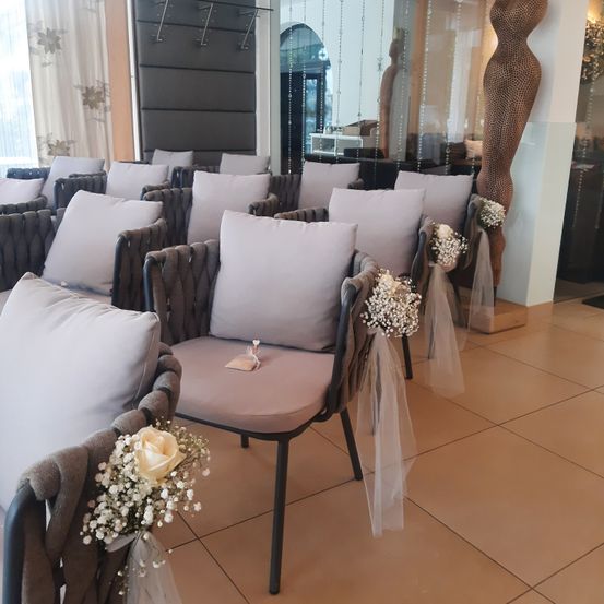 Reihen von gepolsterten Stühlen, geschmückt mit weißen Blumen und Bändern, in einem modernen Raum mit gefliesten Böden und reflektierenden Oberflächen aufgestellt.