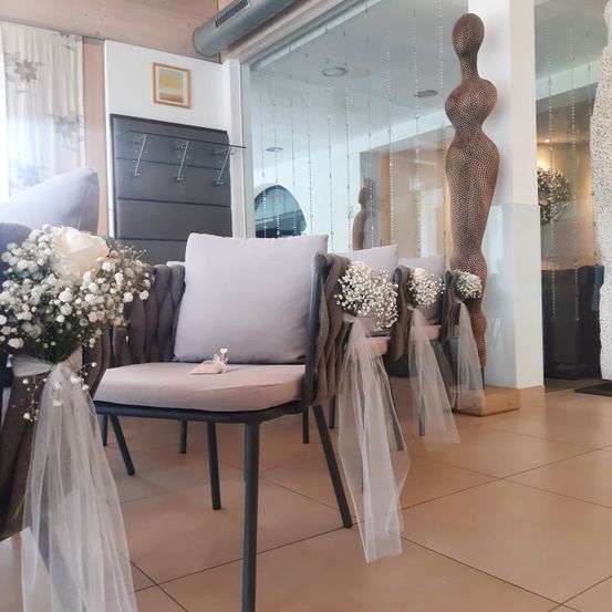 Ein Hochzeitsgang mit Stühlen, die mit weißen Blumen und Stoffbändern geschmückt sind. Eine große dekorative Figur steht im Hintergrund mit Spiegelungen eines Raumes und Glaswänden.