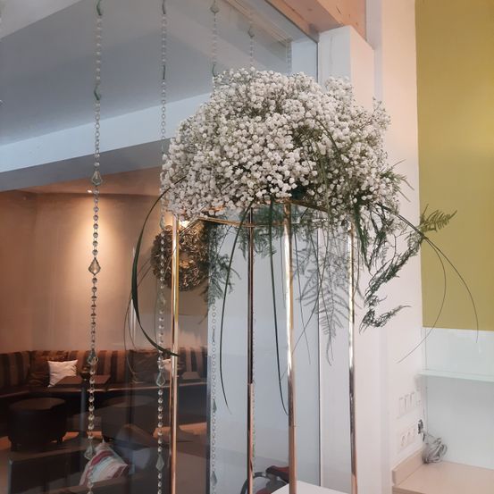 Ein großer Strauß weißer Babys Atemblumen in einem hohen, metallischen Vasen steht in einem modernen Raum. Der Hintergrund zeigt eine gelbe Wand und eine Glastrennwand.