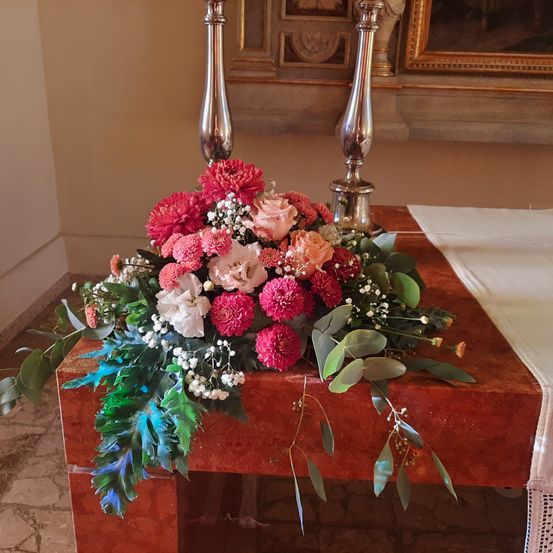 Eine farbenfrohe Blumenarrangement in Rosa, Weiß und Rot schmückt einen hölzernen Altar, ergänzt durch grüne Blätter und silberne Kerzenhalter.