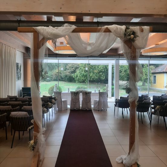Eine Indoor-Hochzeitszeremonie mit einem hölzernen Bogen und weißen Vorhängen, einem langen roten Teppich und Stühlen auf beiden Seiten. Der Raum hat große Glasfenster mit Blick auf eine grasbewachsene Wiese und Bäume.