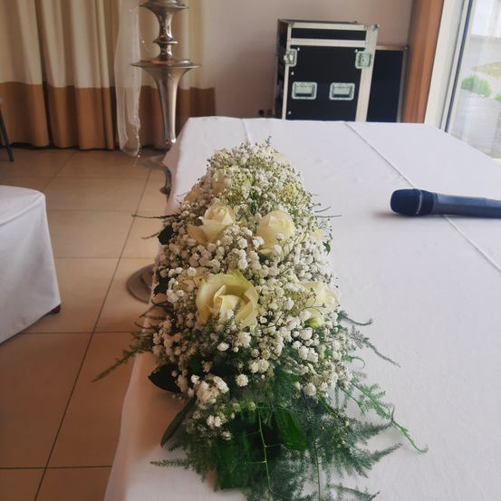 Ein weißer Tisch mit einem Blumenarrangement aus weißen Rosen und Babys Atem. Ein Mikrofon liegt auf dem Tisch, und ein silberner Kerzenständer befindet sich im Hintergrund.