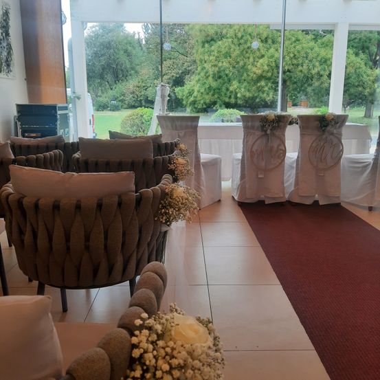 Ein Raum mit Stühlen und Tischen, die mit weißem Stoff bedeckt sind, Blumenarrangements auf den Stühlen und einem roten Teppich auf dem Boden. Draußen befindet sich eine grüne Rasenfläche mit Bäumen.