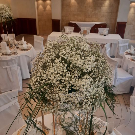 Ein Hochzeitsempfangsbereich mit mehreren Tischen, die mit weißen Tischdecken, Stühlen und einer großen Tischmitte aus weißen Babys-Atem-Blumen gedeckt sind. Jeder Tisch ist mit Tellern, Besteck und Servietten geschmückt. Der Raum hat beige Wände und geflieste Böden.