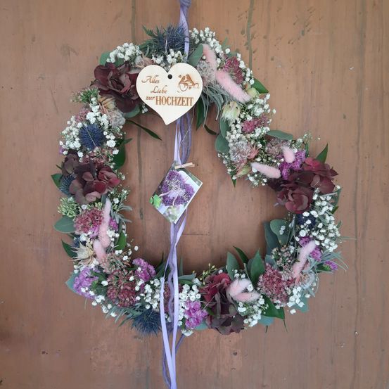 Ein kreisförmiger Blumenkranz mit lila und rosa Blumen und Blättern hängt an einer Holzwand, mit einem herzförmigen Schild, das 'Alles Liebe zur Hochzeit' steht.