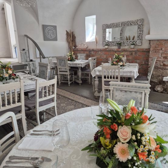 Ein Innenbereich zum Essen mit weißen Möbeln, dekorativen Blumen auf den Tischen und einer Ziegelwand mit einem Spiegel. Eine Kristallleuchte und eine Treppe sind ebenfalls vorhanden.