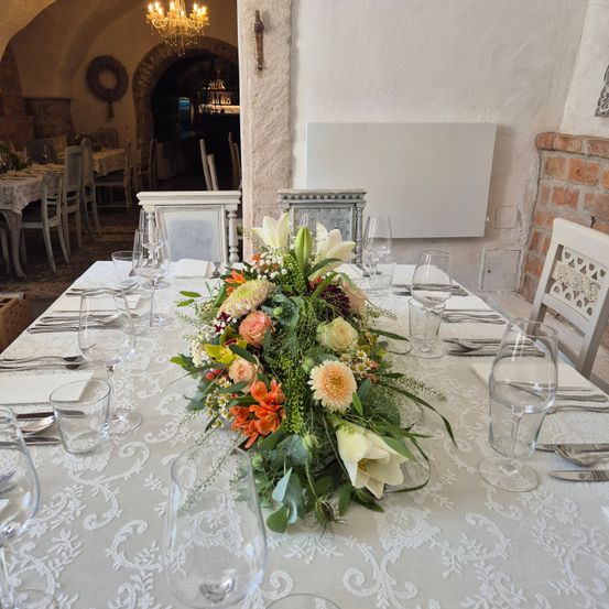 Ein Esstisch ist mit Weingläsern, Tellern und einer großen Blumendekoration gedeckt. Der Tisch ist mit einer weißen Tischdecke bedeckt. Hinter dem Tisch befindet sich eine Wand mit einem weißen Brett und einem Ziegelkamin.