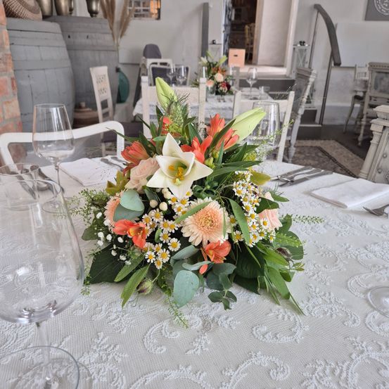 Ein Esstisch mit einem komplexen Blumenarrangement, weißer Tischdecke und Weingläsern. Dahinter ein Esszimmer mit Stühlen und weiteren Tischen.