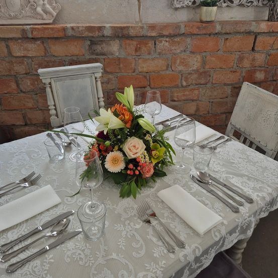 Ein Esstisch mit weißer Spitzentischdecke ist mit Silberwaren, Gläsern und einem Blumenzentrum ausgestattet. Zwei Stühle sind auf beiden Seiten positioniert. Hinter dem Tisch ist eine Ziegelmauer.
