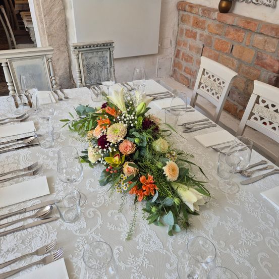 Ein langes Blumenarrangement ziert einen Tisch, der für eine formelle Veranstaltung gedeckt ist. Der Tisch ist von weißen Stühlen umgeben, und die Umgebung umfasst Weingläser, Besteck und eine gemusterte Tischdecke.