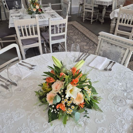 Ein Restaurant mit runden Tischen und Stühlen ist für eine Mahlzeit vorbereitet. Jeder Tisch hat eine Blumenmitte mit Rosen, Lilien und anderen Blumen. Weingläser und Besteck sind ordentlich angeordnet.