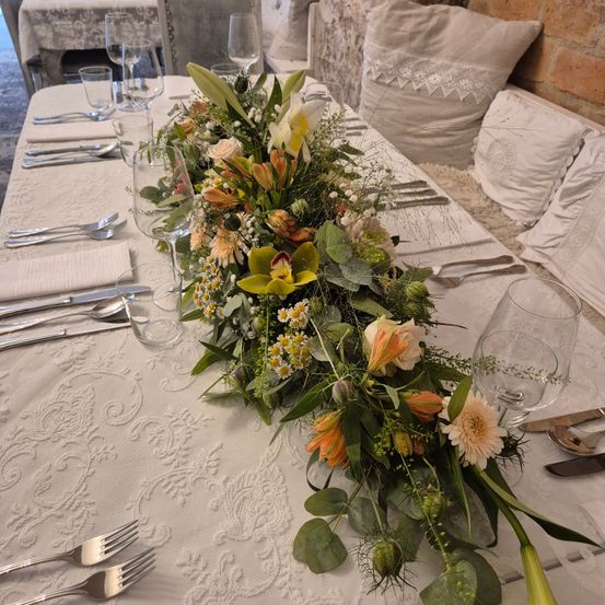 Ein Tisch mit einem aufwendigen Blumen-Zentralstück. Verschiedene Blumen, darunter Rosen und Lilien, sind auf einer weißen Tischdecke arrangiert. Besteck, Gläser und Servietten sind um den Tisch herum platziert.