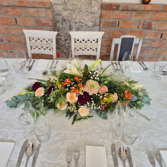 Ein Tisch mit einem Blumenzentrum, weißer Tischdecke und weißen Stühlen ist für eine Mahlzeit vorbereitet. Weingläser, Gabeln, Messer und Löffel sind auf dem Tisch platziert.