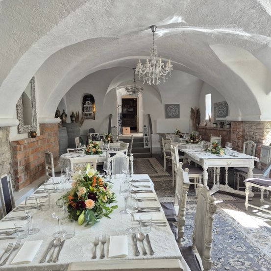 Ein elegantes Esszimmer mit weißen Wänden und gewölbten Decken. Die Tische sind mit Blumen, Weingläsern und Silberbesteck gedeckt. Eine Kristallleuchter hängt von der Decke, und der Raum ist mit weißen Stühlen und Tischen eingerichtet.