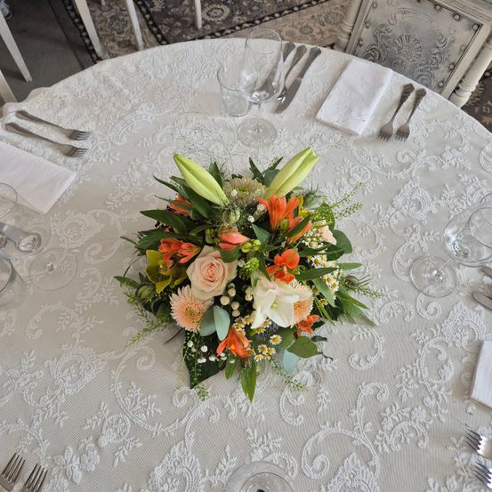 Ein Blumenarrangement auf einem runden Tisch, geschmückt mit einer weißen Spitzentischdecke, umgeben von Besteck und Weingläsern. Das Arrangement besteht aus einer Mischung aus weißen, rosa und orangefarbenen Blumen.