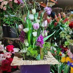Eine Blumenarrangement mit verschiedenen Farben von Blumen, einschließlich rosa, lila und weiß, wird in einer lila Box mit einem natürlichen Weidendesign ausgestellt. Es ist von anderen Blumen und Pflanzen umgeben.