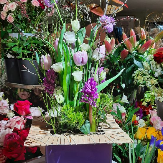 Eine Blumenarrangement mit verschiedenen Farben von Blumen, einschließlich rosa, lila und weiß, wird in einer lila Box mit einem natürlichen Weidendesign ausgestellt. Es ist von anderen Blumen und Pflanzen umgeben.