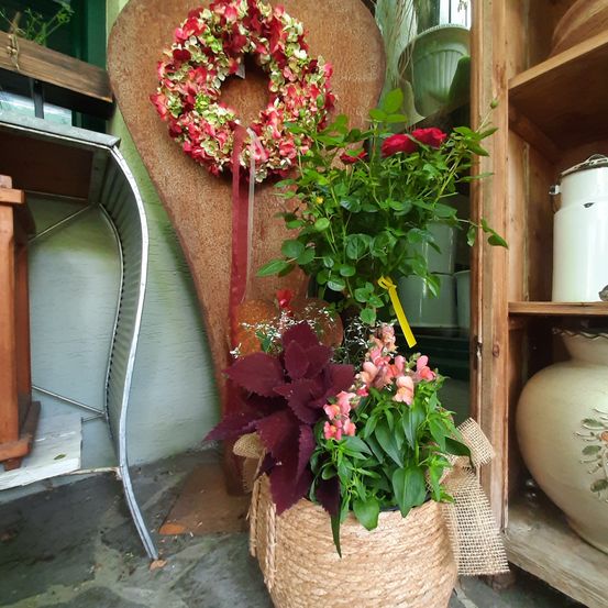 Ein dekorativer Korb mit roten und rosa Blumen steht vor einer Holzwand, die mit einem roten Kranz geschmückt ist. In der Nähe befindet sich ein Metallstuhl und ein Holzregal mit verschiedenen Gegenständen.